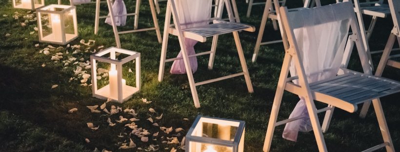 Multiple white chairs draped in white tool are sitting in a row ready for a wedding with flower petals and candle lanterns down the aisle.