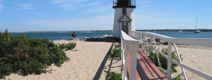 There is a lighthouse on a beach in Nantucket with a walkway on the beach leading to it.