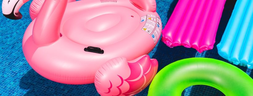 A pink flamingo inflatable pool floatie floats in a pool along with a pink and a blue lounger and a green ring.